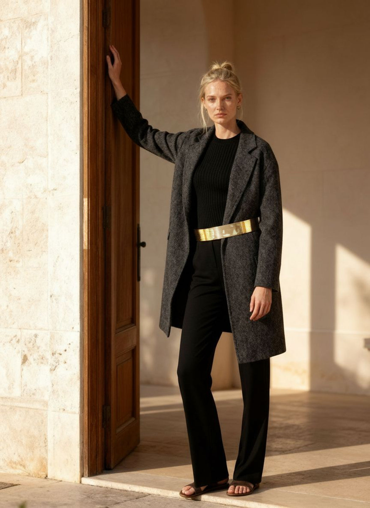 Woman resting a hand on a stone doorway in an open charcoal wool coat and black knit, the gold coil belt at her waist.
