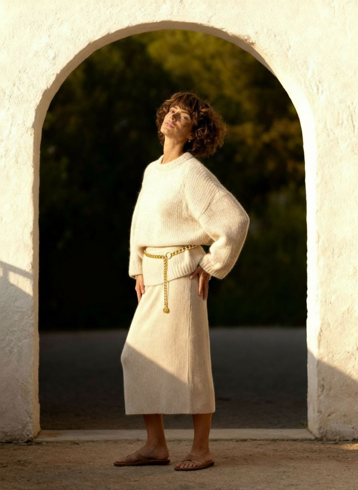 Woman standing in a plaster archway in an oatmeal knit set, the gold drop chain at her waist.