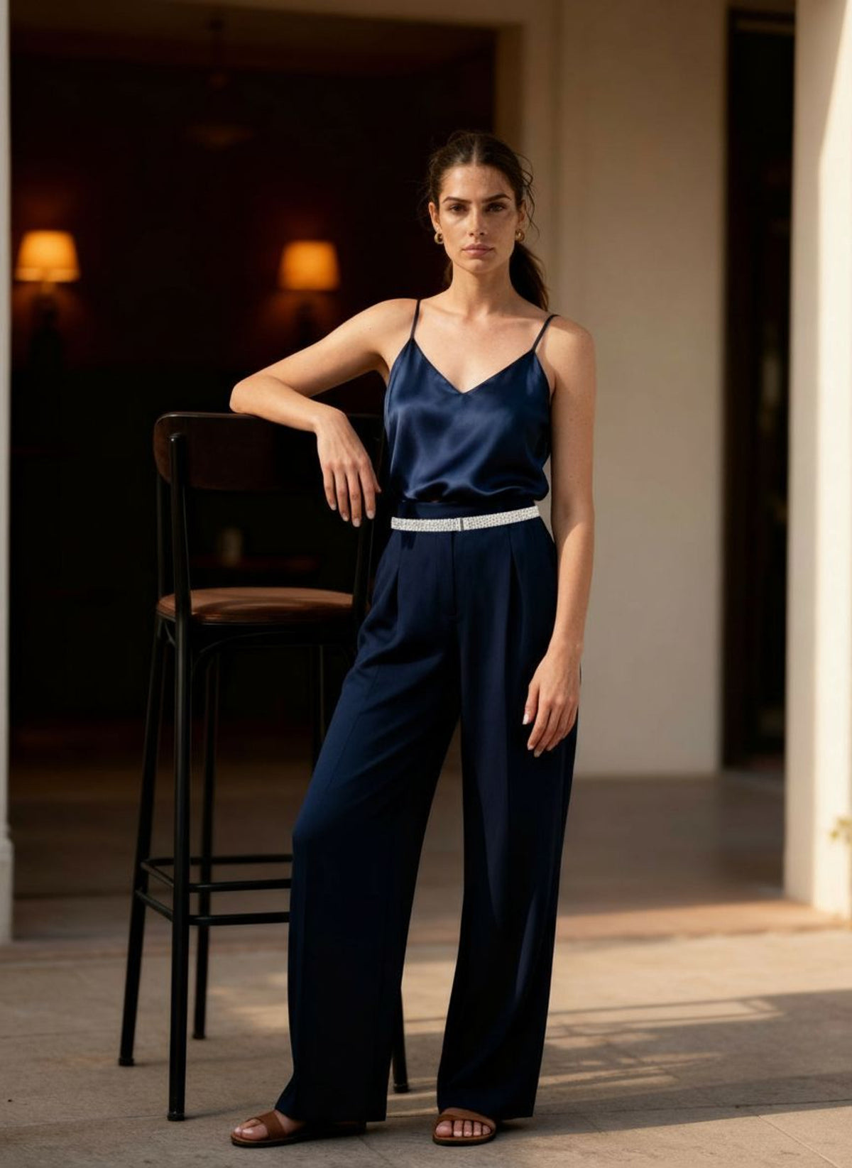 Woman leaning on a bar stool in a navy silk camisole and trousers, the pave crystal belt at her waist.